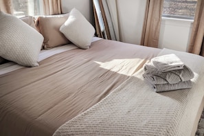 Una cama bien hecha con cubrecama color beige y almohadas blancas, una toalla blanca doblada y una ventana con cortinas.
