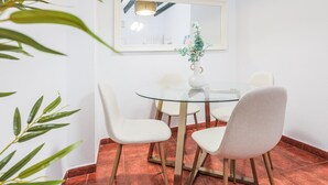 Un coin repas comprenant une table ronde en verre, quatre chaises blanches, un vase blanc contenant des plantes vertes et un luminaire suspendu.