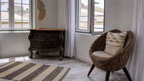 Una habitación con un ventanal grande, una silla de mimbre con cojín, una pequeña mesa de madera y una hoja decorativa en la pared.