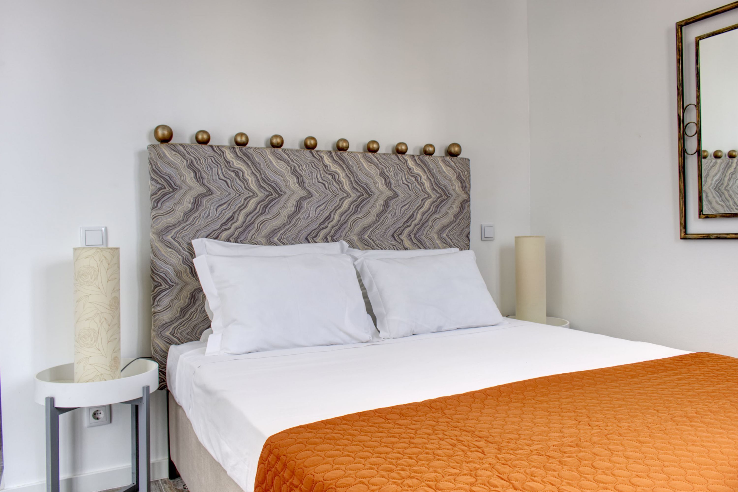 Una cama bien tendida con ropa de cama blanca y naranja, un cabecero con estampado de mármol y dos lámparas de noche.