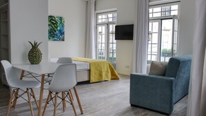 A modern living room with a sofa, a dining table with chairs, a TV mounted on the wall, and a window with curtains. - PB STD Mar Timo - Palacete da Baixa (Faro)