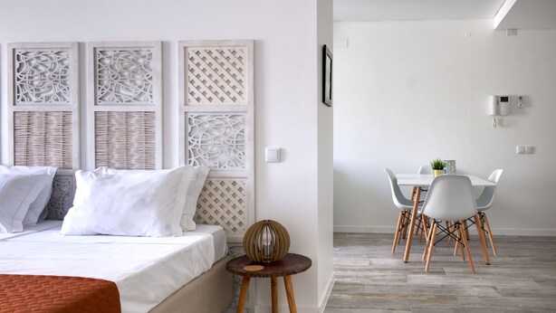 A hotel room with a bed, a small round table with a decorative lamp, a dining area with a white table and chairs, and a wall-mounted light fixture. - PB STD Mar Timo - Palacete da Baixa (Faro)