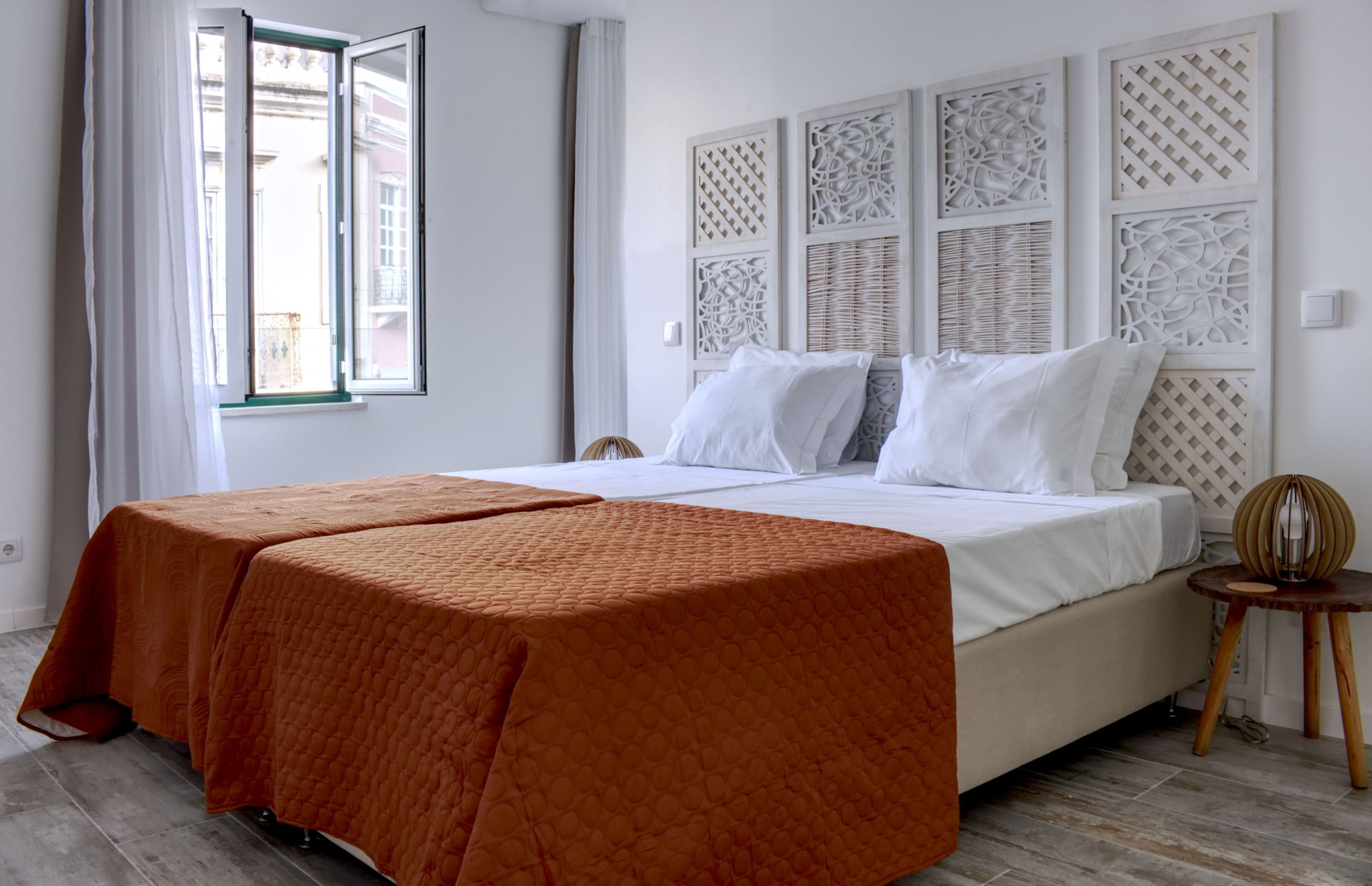 A double bed with white linens, a wooden headboard with intricate carvings, and a view of the outside through an open window.