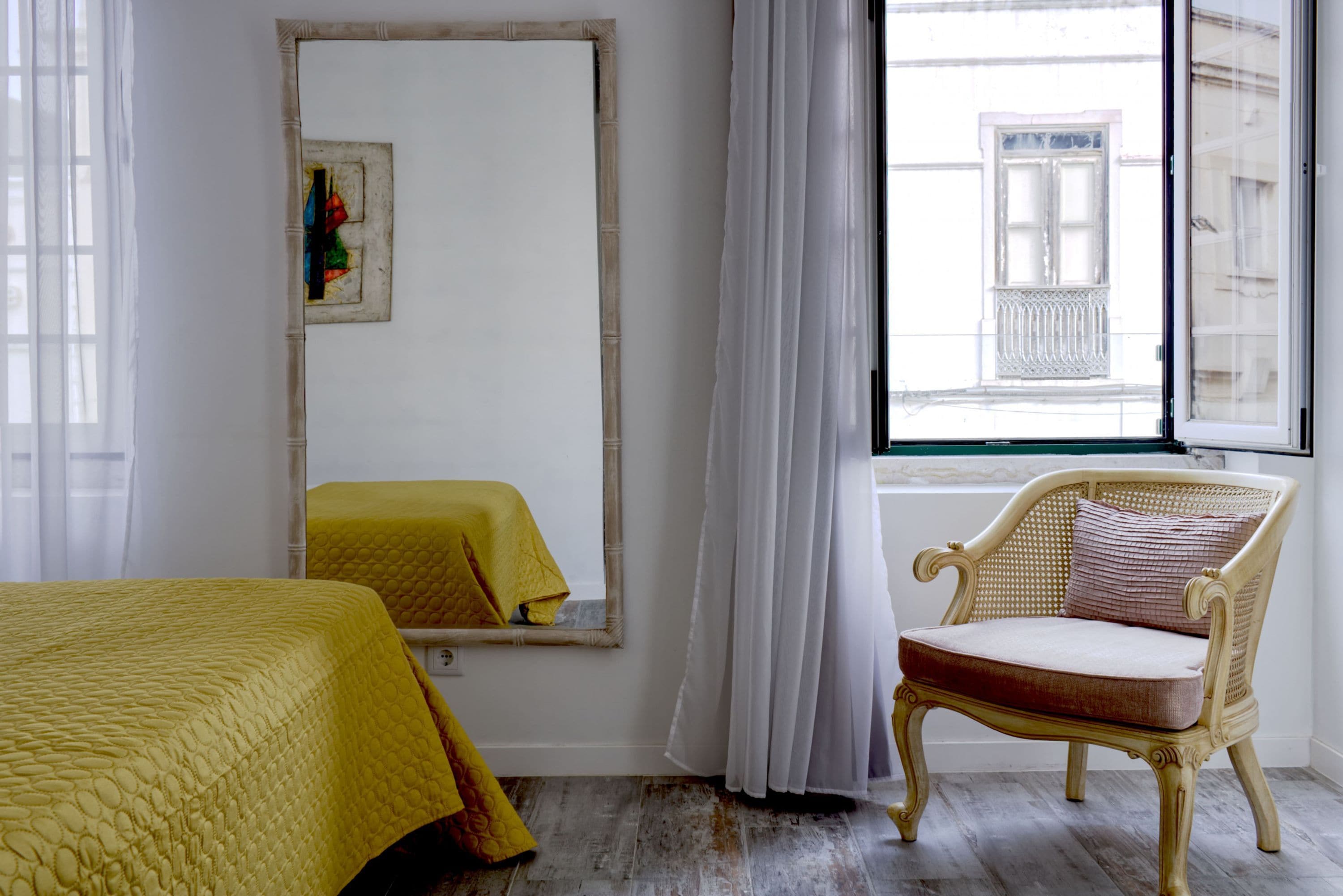 Ein Zimmer mit einem Bett, einem Sessel, einem großen Spiegel und einem Fenster mit Blick auf Gebäude.
