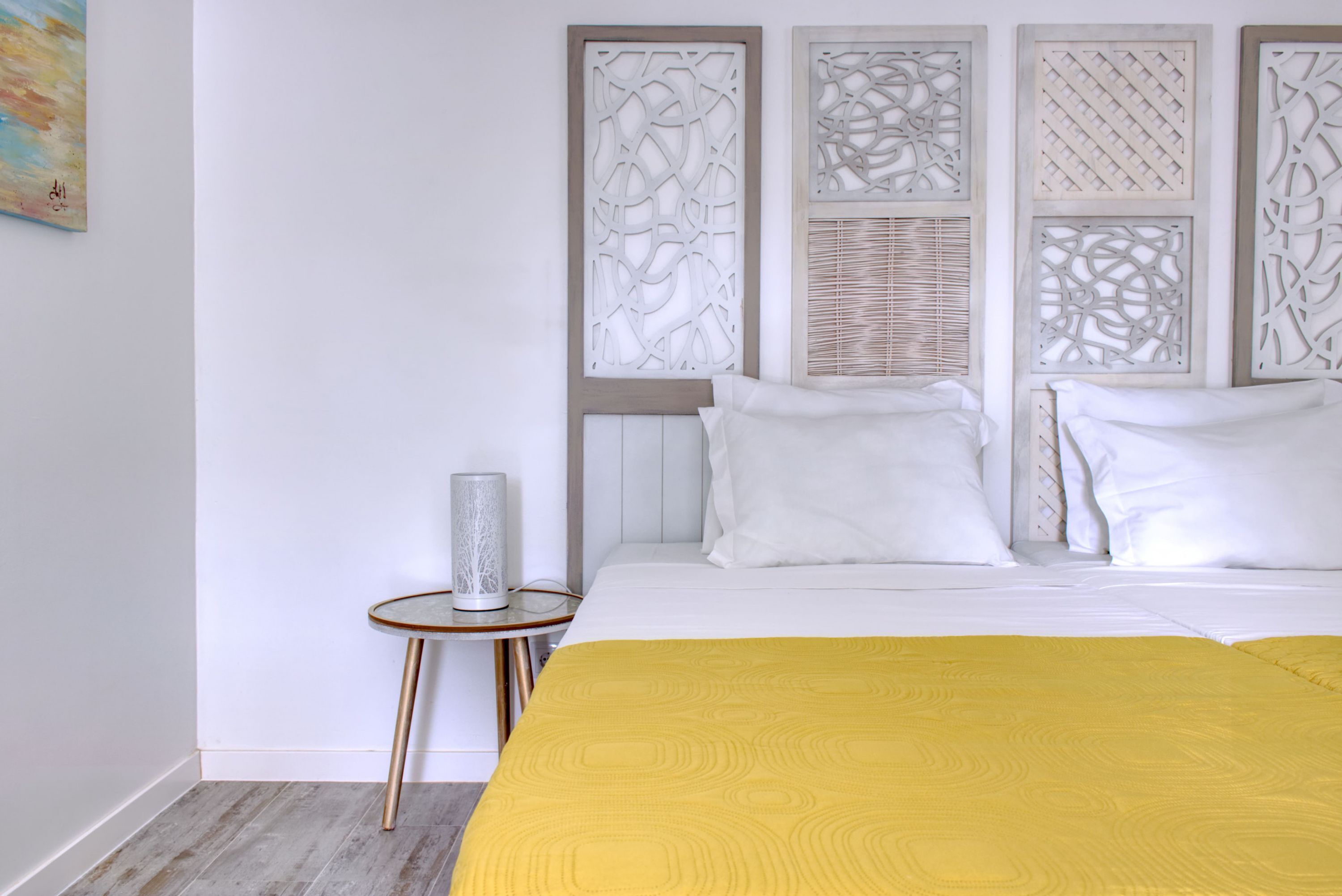 Une chambre à coucher avec un lit, du linge de lit blanc, un plaid jaune, une table de chevet avec une lampe et des panneaux décoratifs sur le mur.