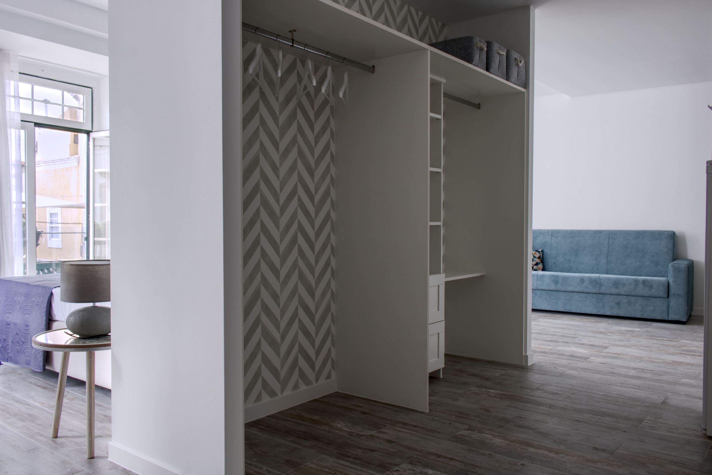 Een kamer met een kledingkast, een blauwe bank en een klein tafeltje met een lamp.