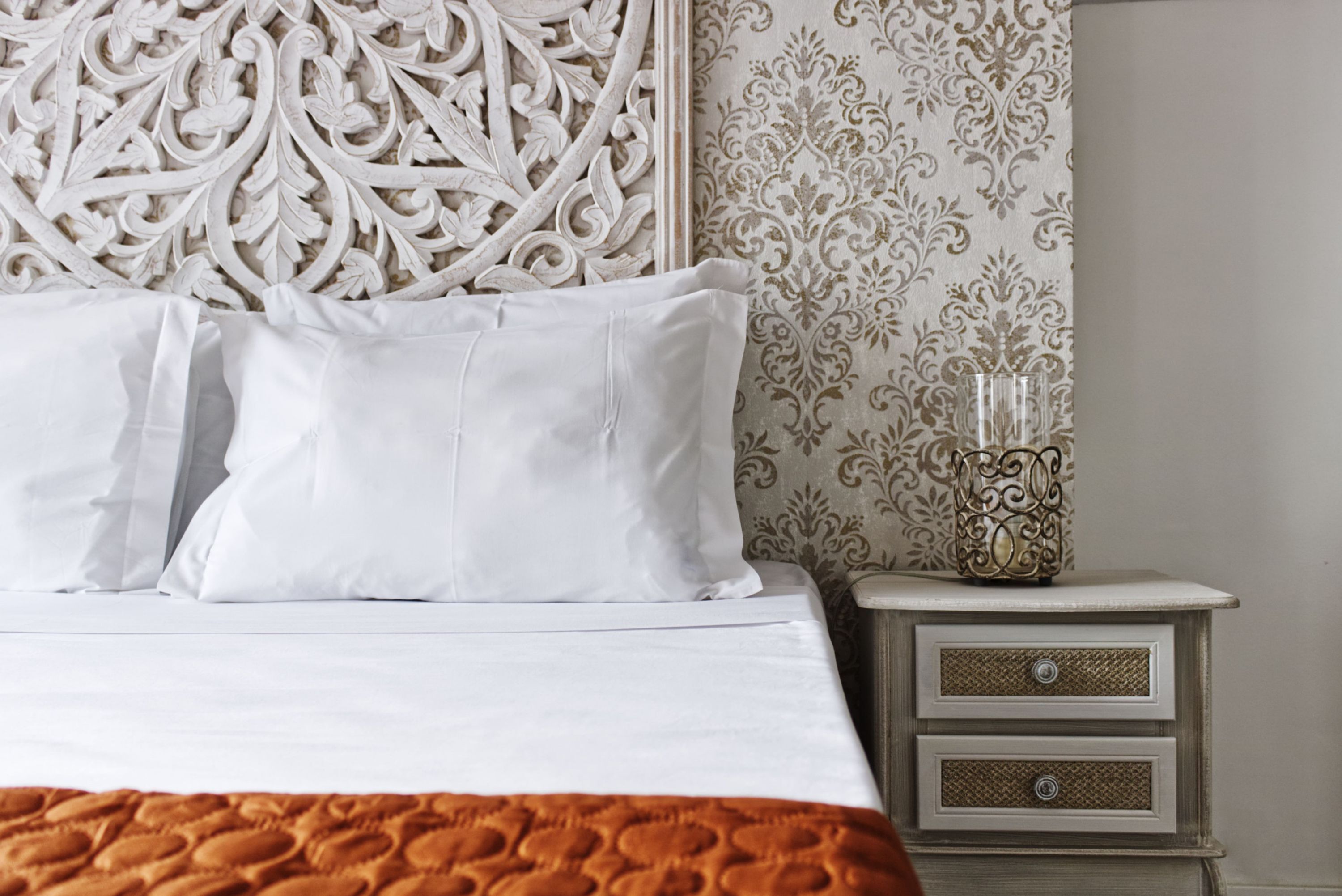 A neatly made bed with white linens and a decorative headboard, a bedside table with a vase, and a patterned wallpaper.