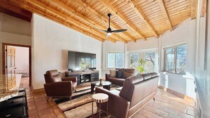 Ein geräumiges Wohnzimmer mit Holzbalkendecke, einem Flachbildfernseher, Ledersofas und einem Essbereich mit Tisch und Stühlen.