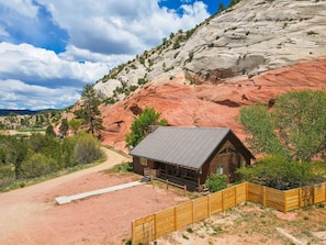 A rustic cabin with a metal roof, surrounded by a wooden fence, set against a backdrop of red rock formations and sparse vegetation.