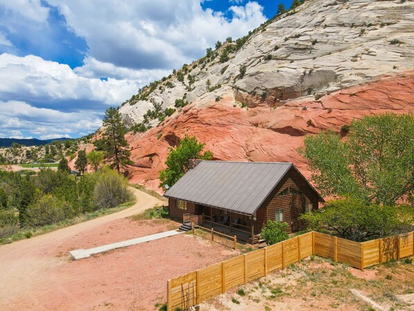 A rustic cabin with a metal roof, surrounded by a wooden fence, set against a backdrop of red rock formations and sparse vegetation.