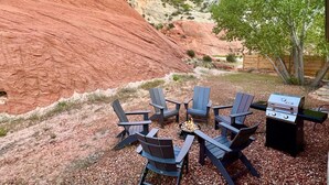 An outdoor seating area with Adirondack chairs and a grill, set against a backdrop of red rock formations and a tree.