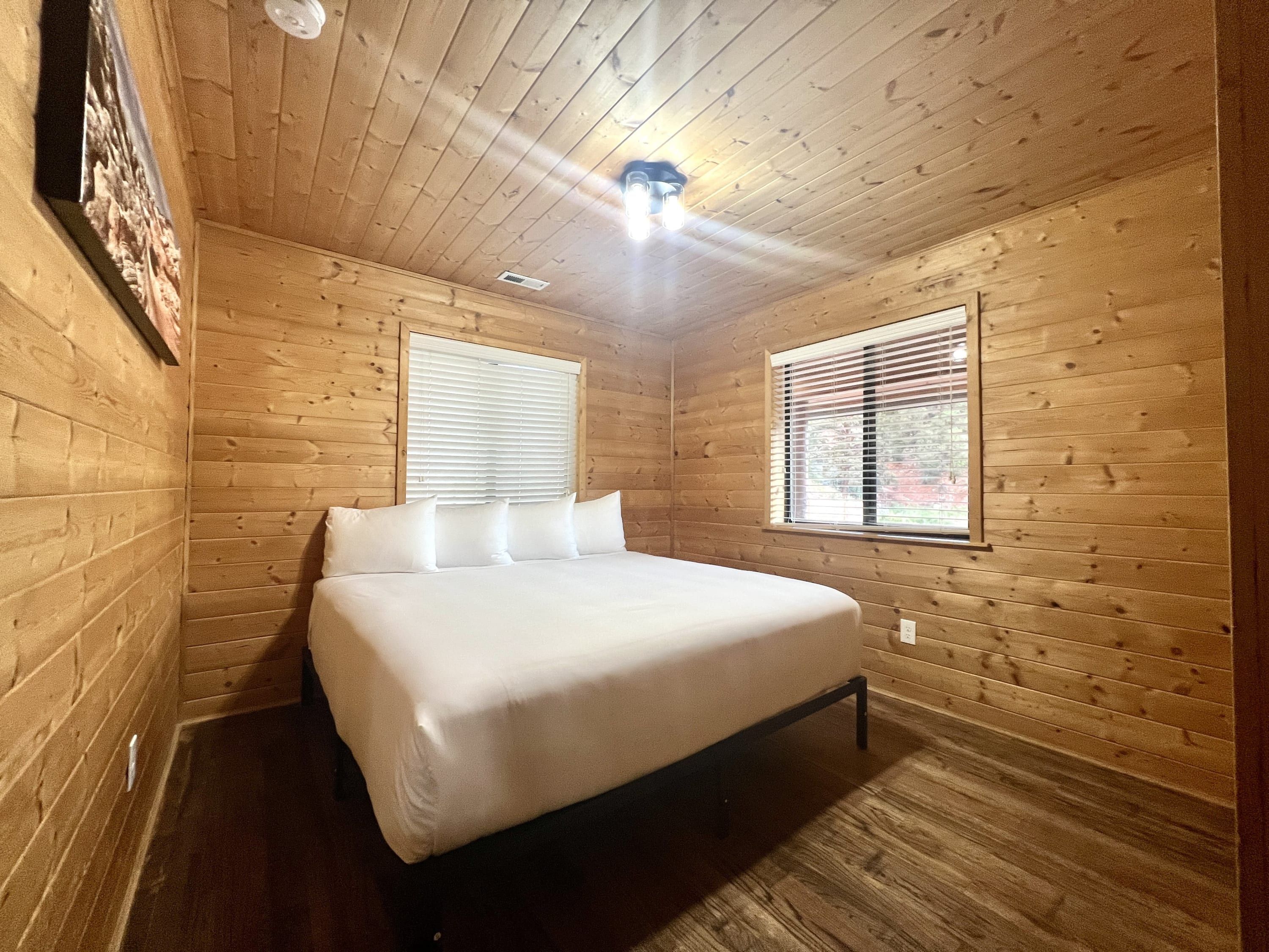Ein Zimmer mit Holzwänden, einem Bett, zwei Fenstern und einer Deckenleuchte.