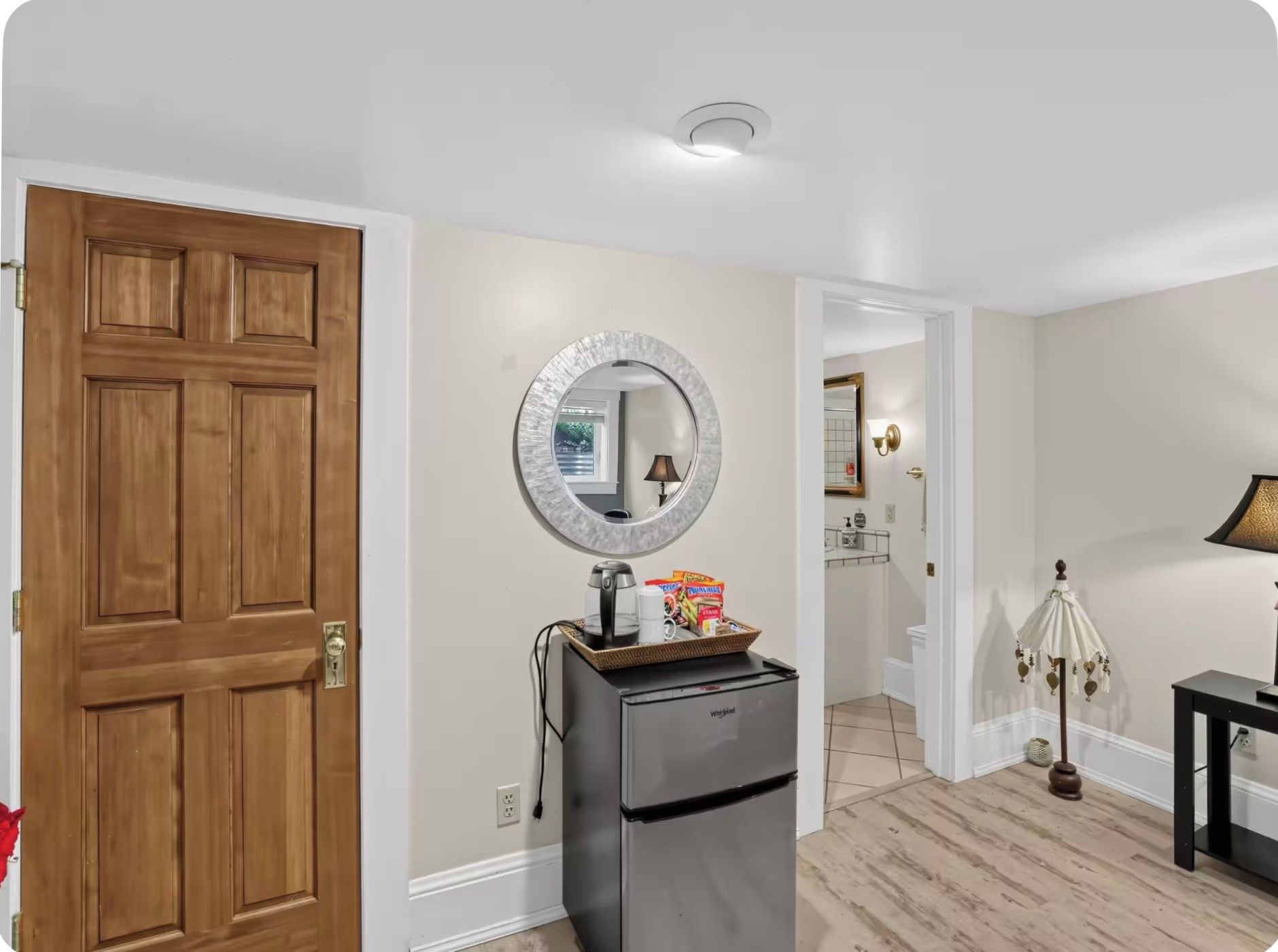 A hallway with a wooden door, a refrigerator with a basket of items, a round mirror, and a lamp.