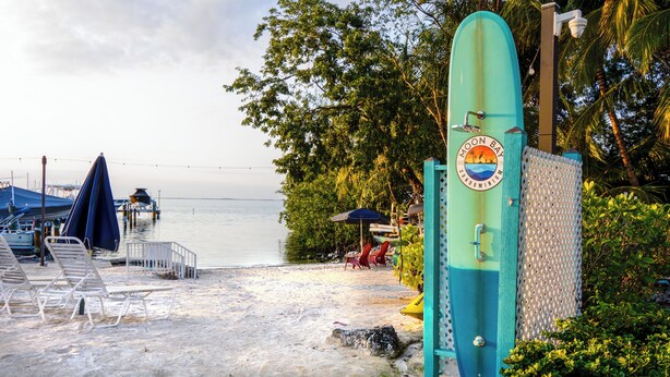 Une plage avec une porte turquoise sur laquelle est inscrit « Moon Bay Condominium », une jetée avec des bateaux et des chaises de plage.