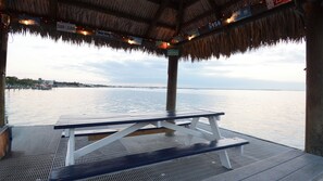 A covered outdoor seating area with a table and benches, overlooking a body of water with a distant shoreline.