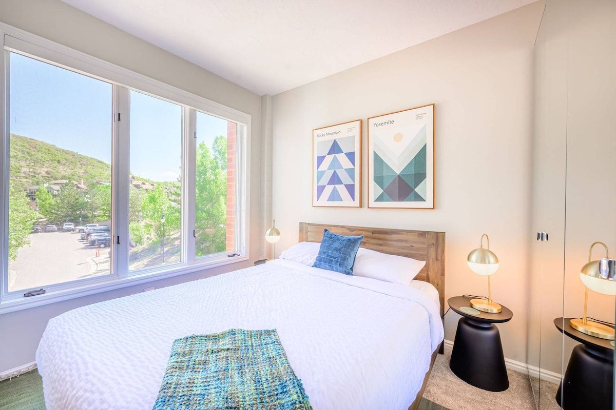 A hotel room with a large bed, two bedside tables, a window with a view of a parking lot, and two framed artworks on the wall.
