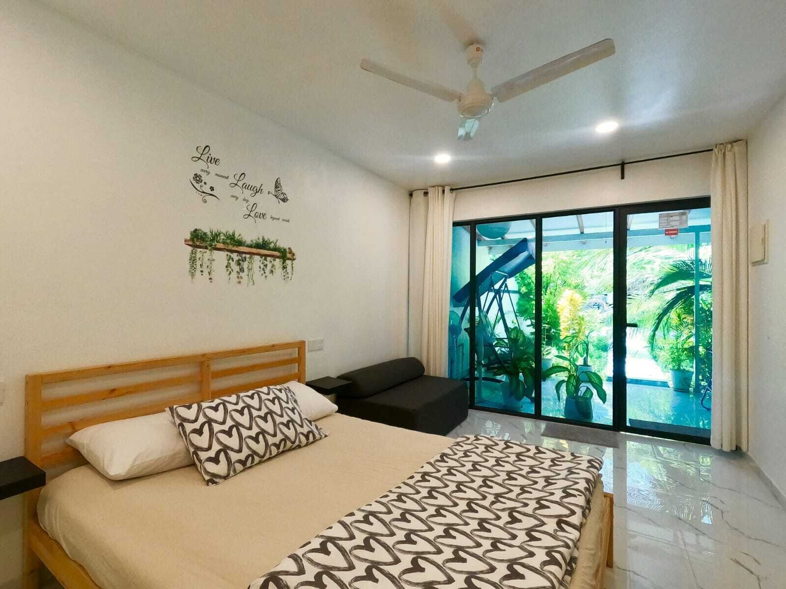 Un dormitorio con cama, ventilador de techo, ventana con vistas a la vegetación y una pared con una frase decorativa.