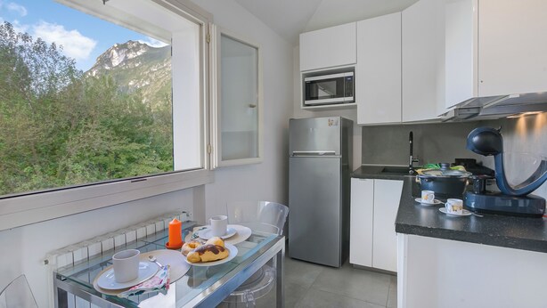 A modern kitchen with a refrigerator, microwave, and stove, a glass dining table with plates and cups, and a window with a mountain view. - Porto Letizia Mimosa 6, Porlezza, Italy (Porlezza)