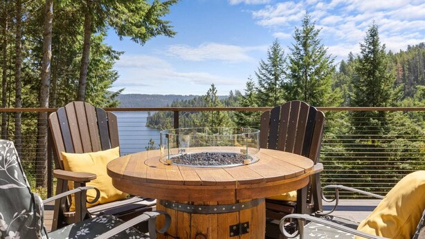 A wooden table with a glass enclosure and a hot surface, surrounded by Adirondack chairs with yellow cushions, overlooking a lake and forest. - Cave Bay Escape - Stunning CDA Lake View, Deck, Grill, Blackstone, Fire Pit, and near Boat Launch! (Worley)