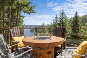 A wooden table with a glass enclosure and a hot surface, surrounded by Adirondack chairs with yellow cushions, overlooking a lake and forest. - Cave Bay Escape - Stunning CDA Lake View, Deck, Grill, Blackstone, Fire Pit, and near Boat Launch! (Worley)