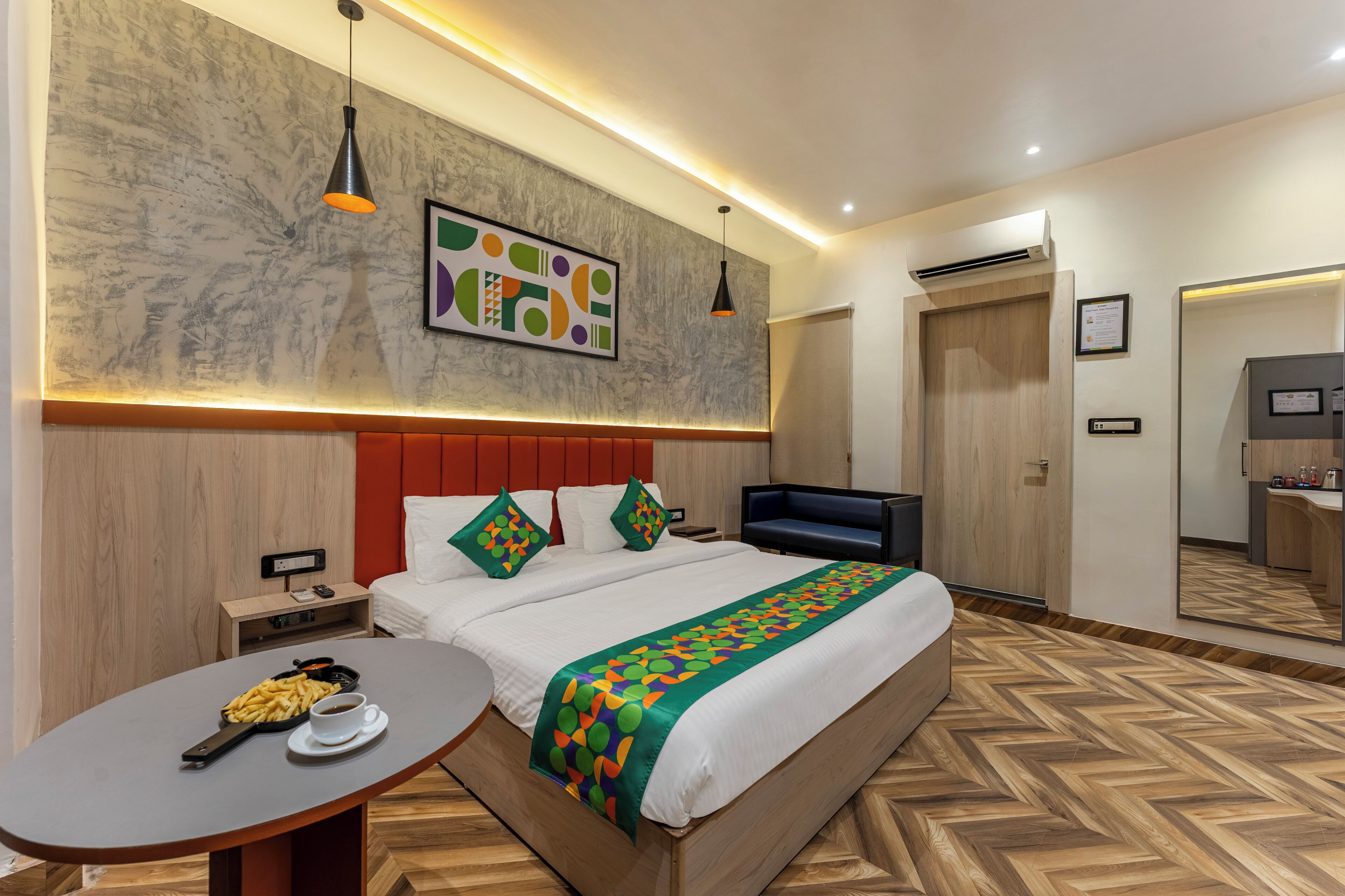 A modern hotel room with a large bed, a desk with a laptop, a small table with a cup and a plate of food, and a large mirror.