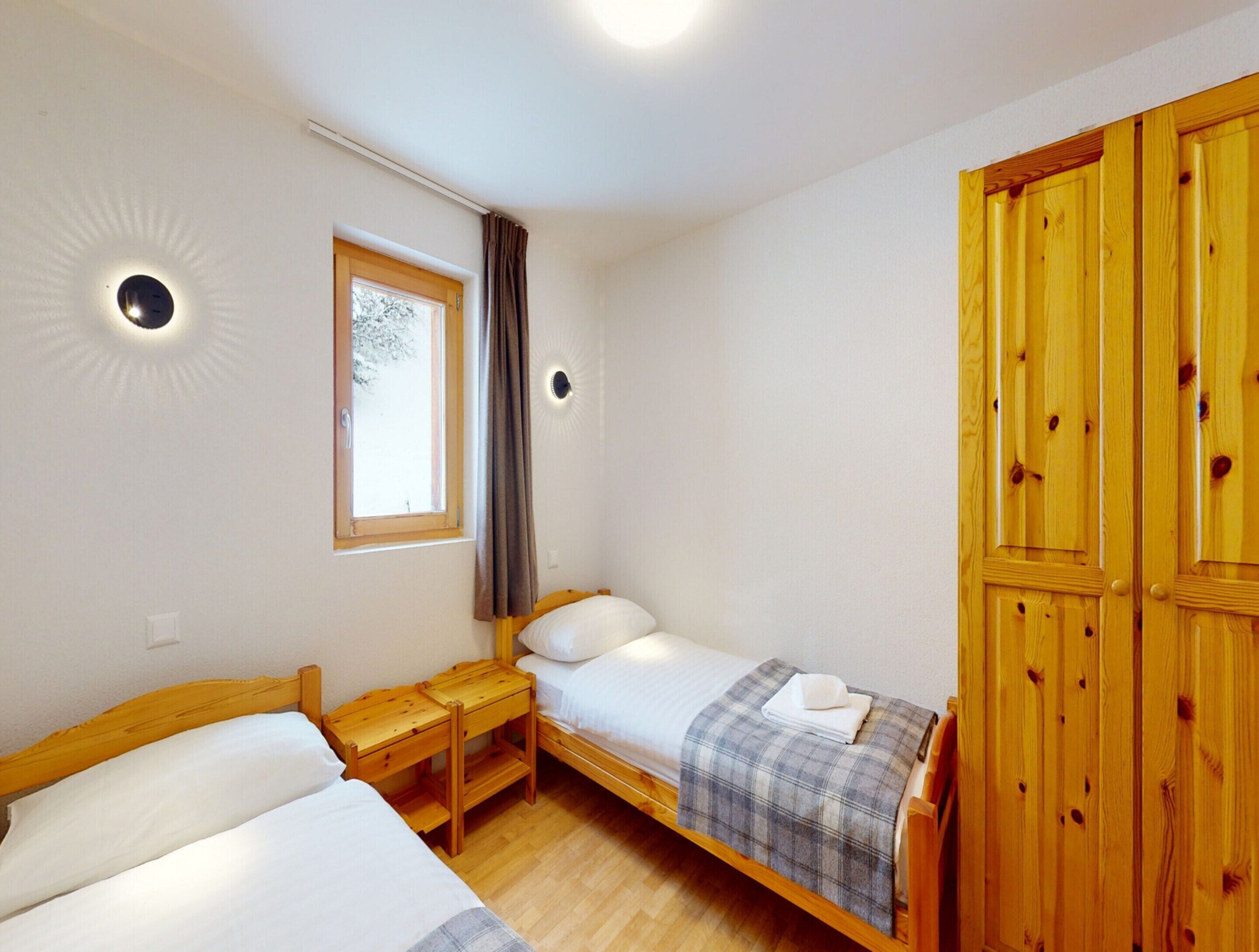 Ein Schlafzimmer mit zwei Einzelbetten, einem Fenster mit Vorhängen, einem Holzschrank und einem Nachttisch mit Lampe.