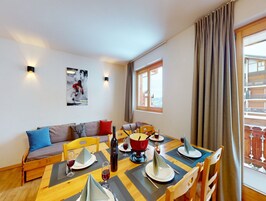 A dining area with a wooden table set for four, a sofa with cushions, and a window with curtains.