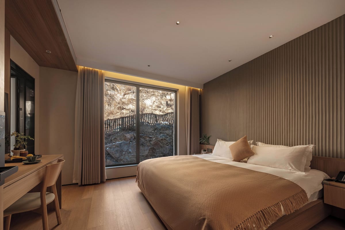 A modern bedroom with a large bed, a desk, and a view of a rocky outdoor area through a large window.