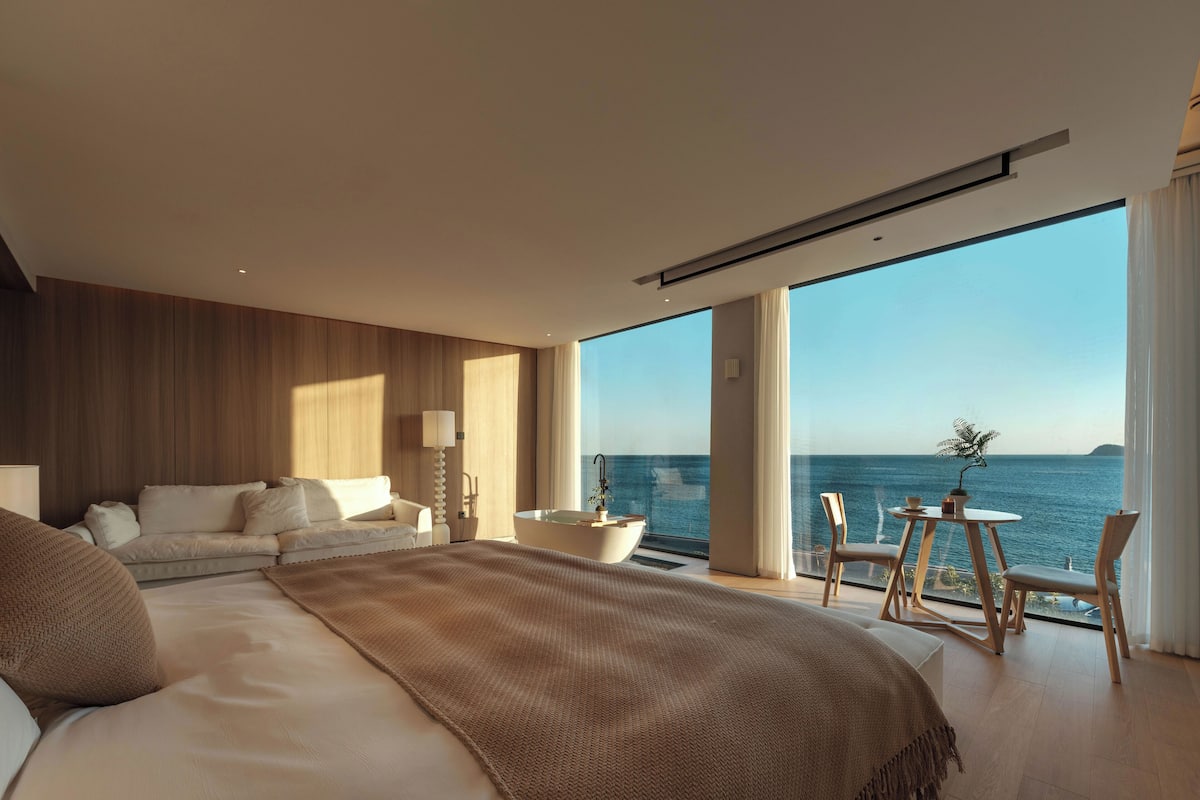 A modern bedroom with a large bed, a view of the sea, and a small table with chairs.