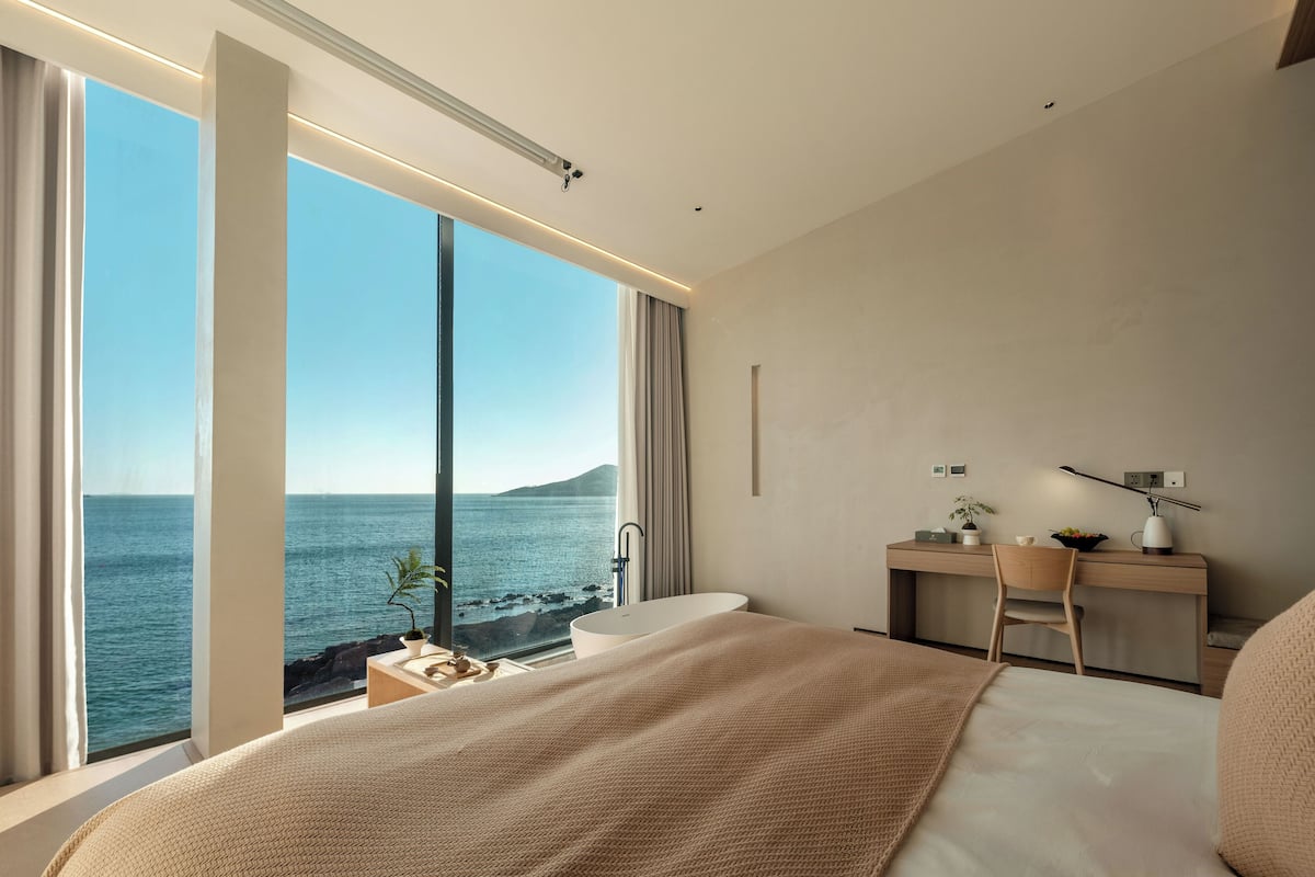 A modern bedroom with a large window overlooking the sea, a bed with a beige blanket, a desk with a chair, and a small plant.