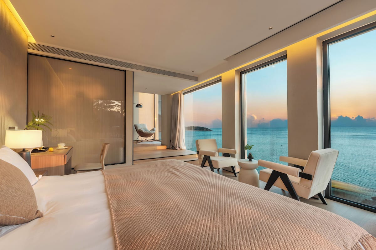 A modern hotel room with a large bed, a desk, and a seating area with a view of the sea.