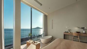 Un dormitorio moderno con un amplio ventanal con vista al mar, una bañera integrada y una zona de escritorio minimalista.