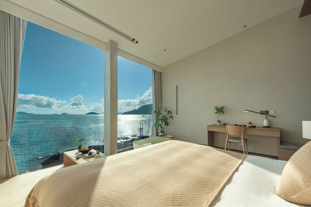 A modern hotel room with a large bed, a desk, and a view of the sea.