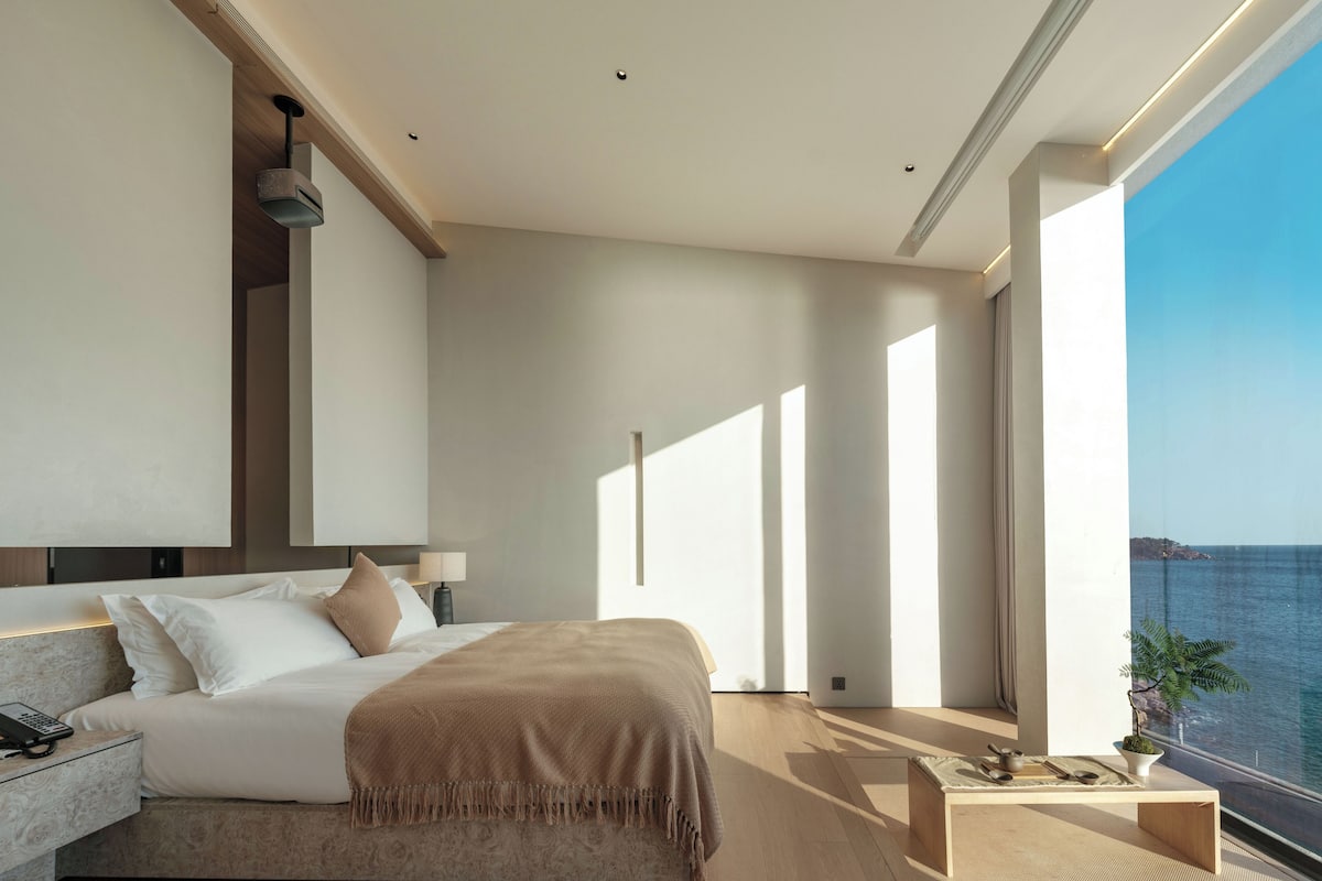 A modern bedroom with a large bed, bedside table, and a view of the sea.