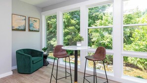 A room with a green sofa, two bar stools, a round table, and large windows with a view of trees. - Magnolia Deluxe King Studio at The Dogwood (Maggie Valley)