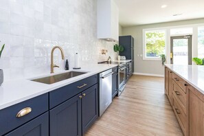 Una cocina moderna con armarios oscuros, encimera blanca, electrodomésticos de acero inoxidable y un amplio ventanal que proporciona luz natural.