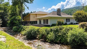 A two-story house with a front porch, a stone chimney, and a small front yard. - Chic Creekside Bungalow at The Dogwood (Maggie Valley)