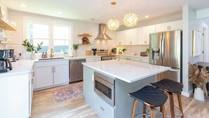 A modern kitchen with white cabinetry, stainless steel appliances, and a large island with bar stools. - Chic Creekside Bungalow at The Dogwood (Maggie Valley)
