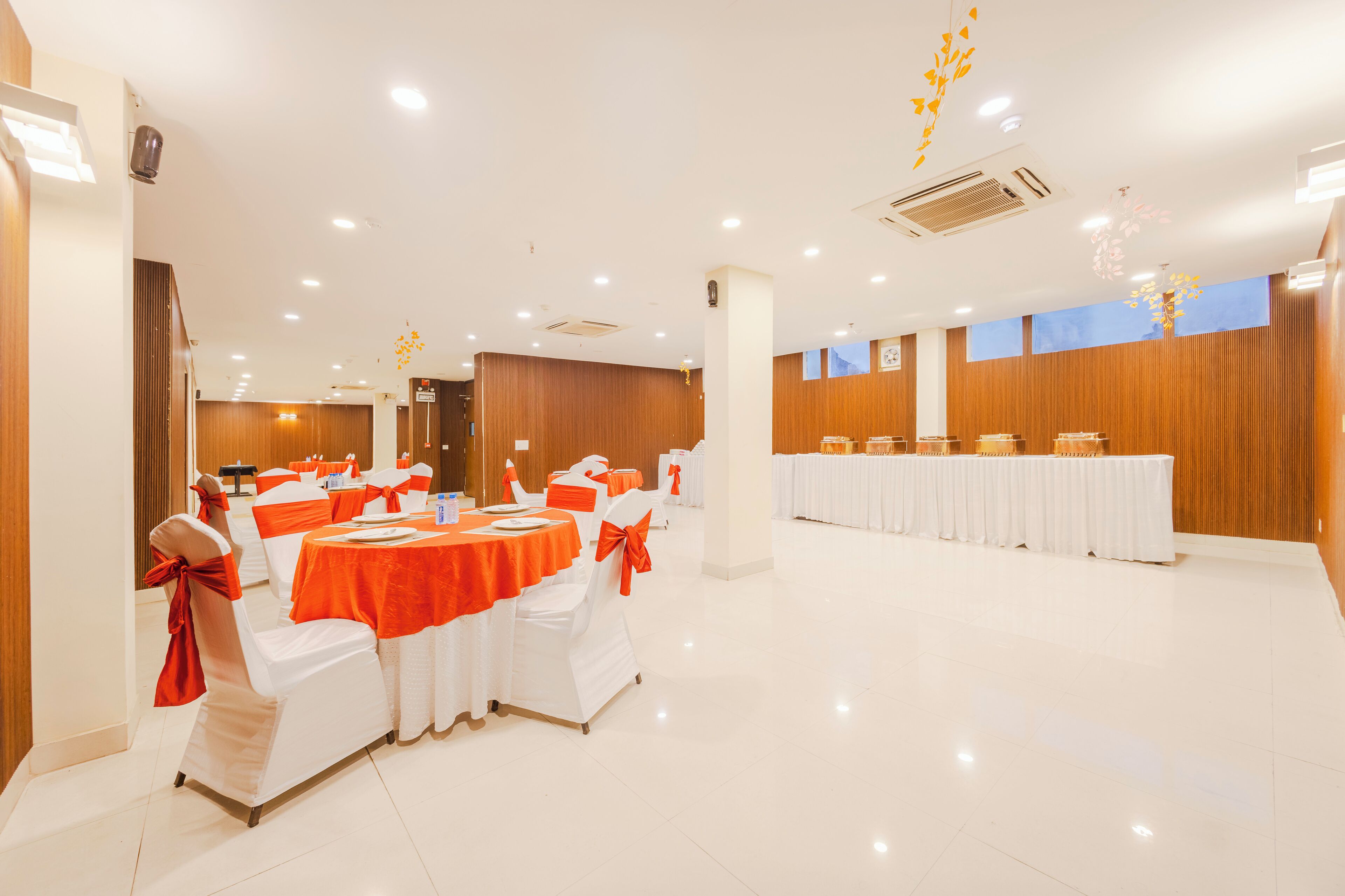 A spacious banquet hall with round tables and white chairs, featuring a modern design with wooden paneling and recessed lighting.