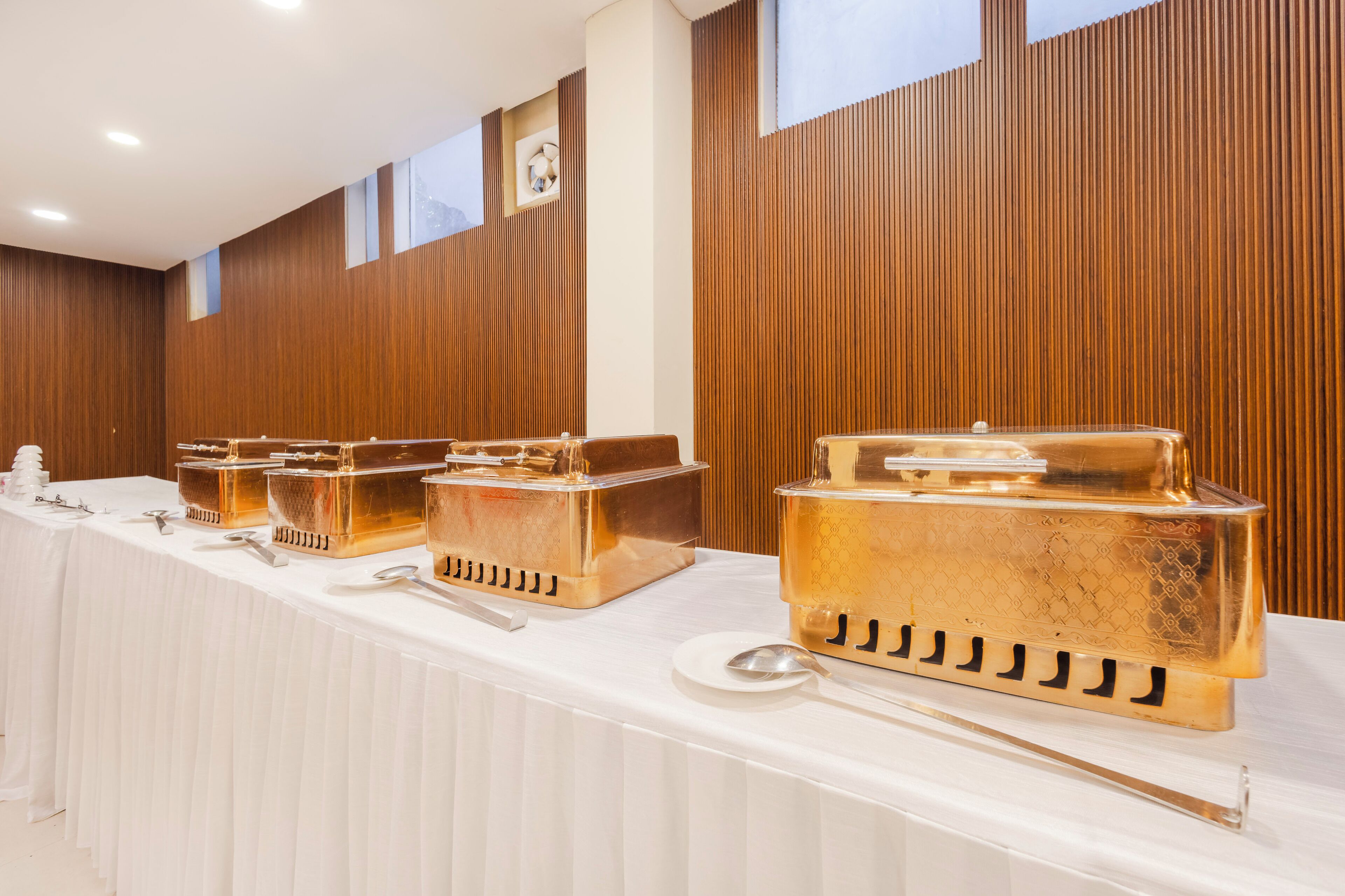 A long table with gold-plated serving trays and utensils, set against a wooden wall with vertical panels and a window.