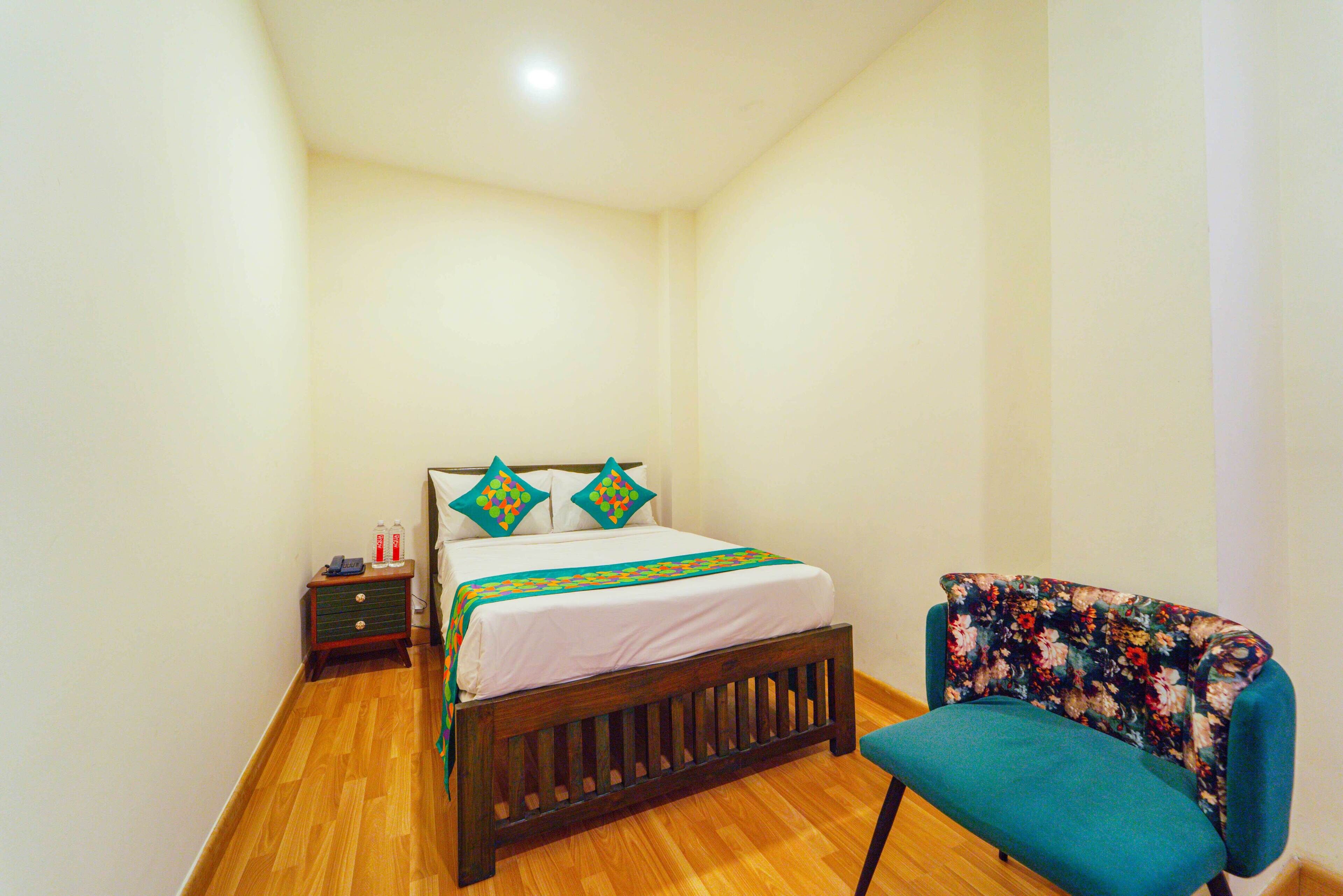 A bedroom with a wooden floor, a bed with a white bedspread and two decorative pillows, a small bedside table with a lamp, and a floral-patterned chair.