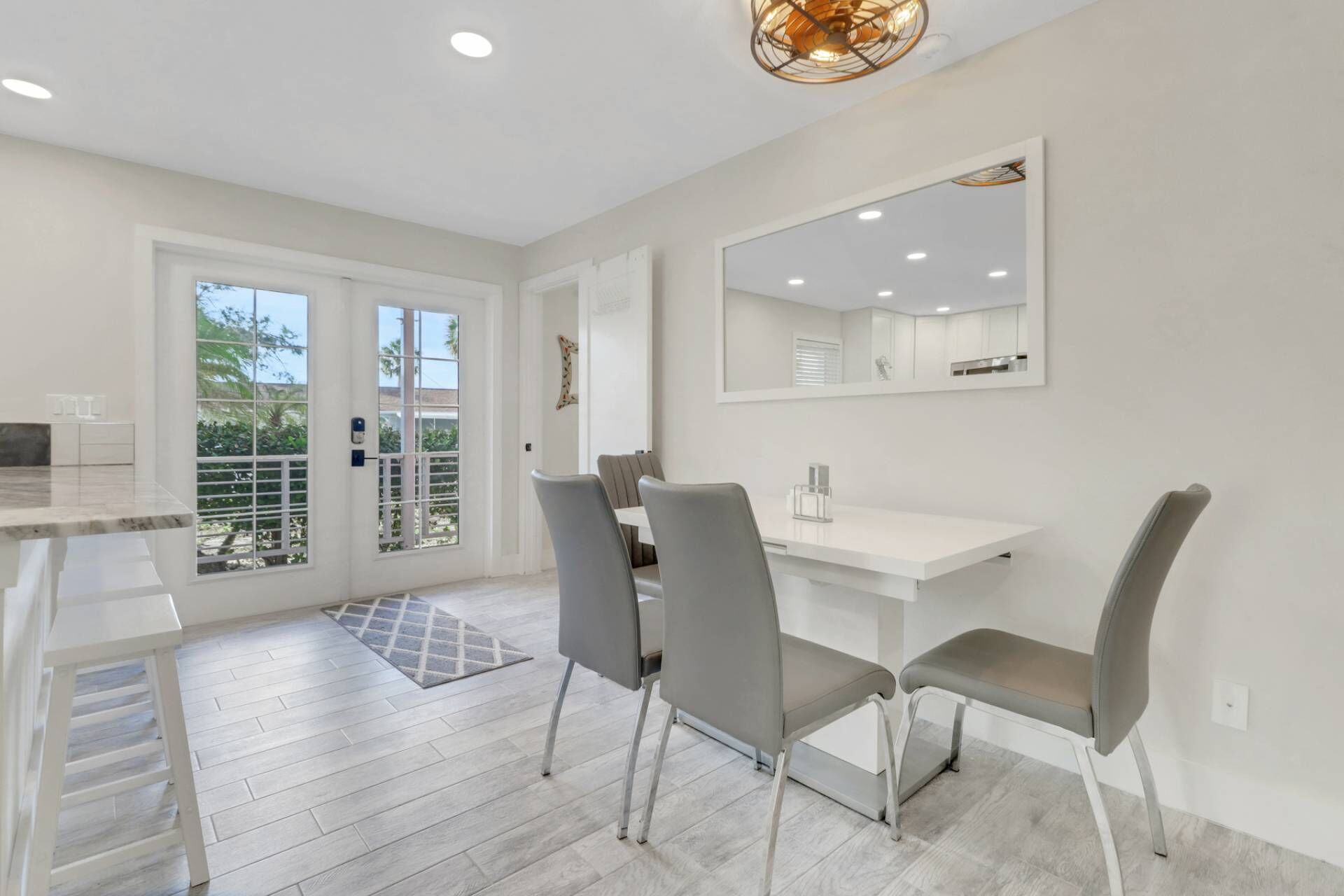 A modern kitchen with white cabinetry, a large window, and a dining area with grey chairs and a white table.