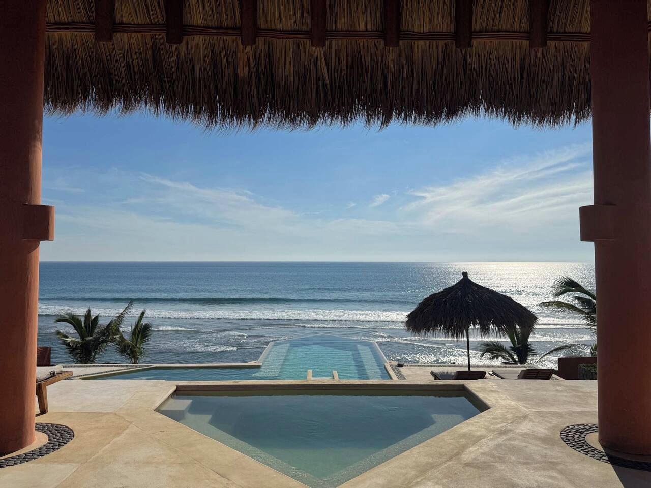 An infinity pool with a view of the ocean, a thatched roof structure, and palm trees.