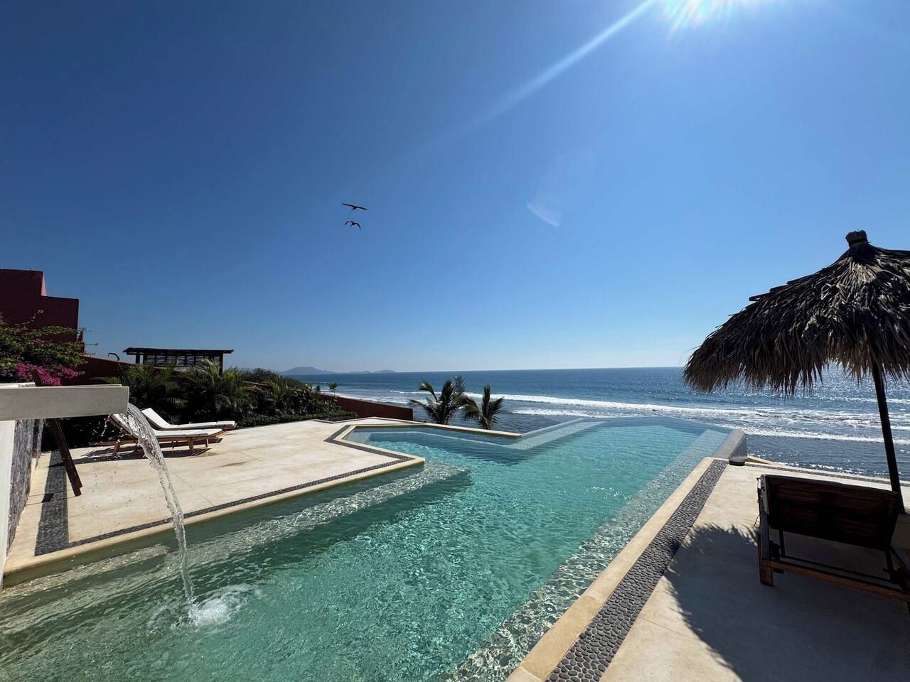 Una piscina con vistas al océano, sillas de descanso y una sombrilla de paja.