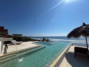 A pool with a view of the ocean, lounge chairs, and a thatched umbrella.