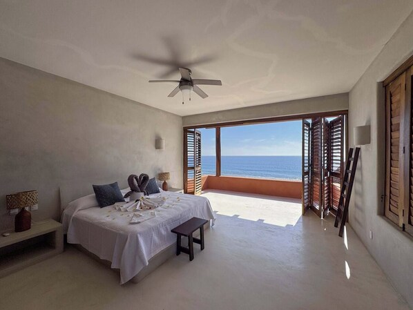 A bedroom with a bed, bedside table, lamp, and a view of the ocean.