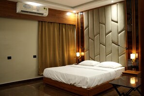A hotel room with a wooden bed, beige curtains, a wall-mounted air conditioner, and a nightstand with a lamp. - Banmali regency (Golmuri)