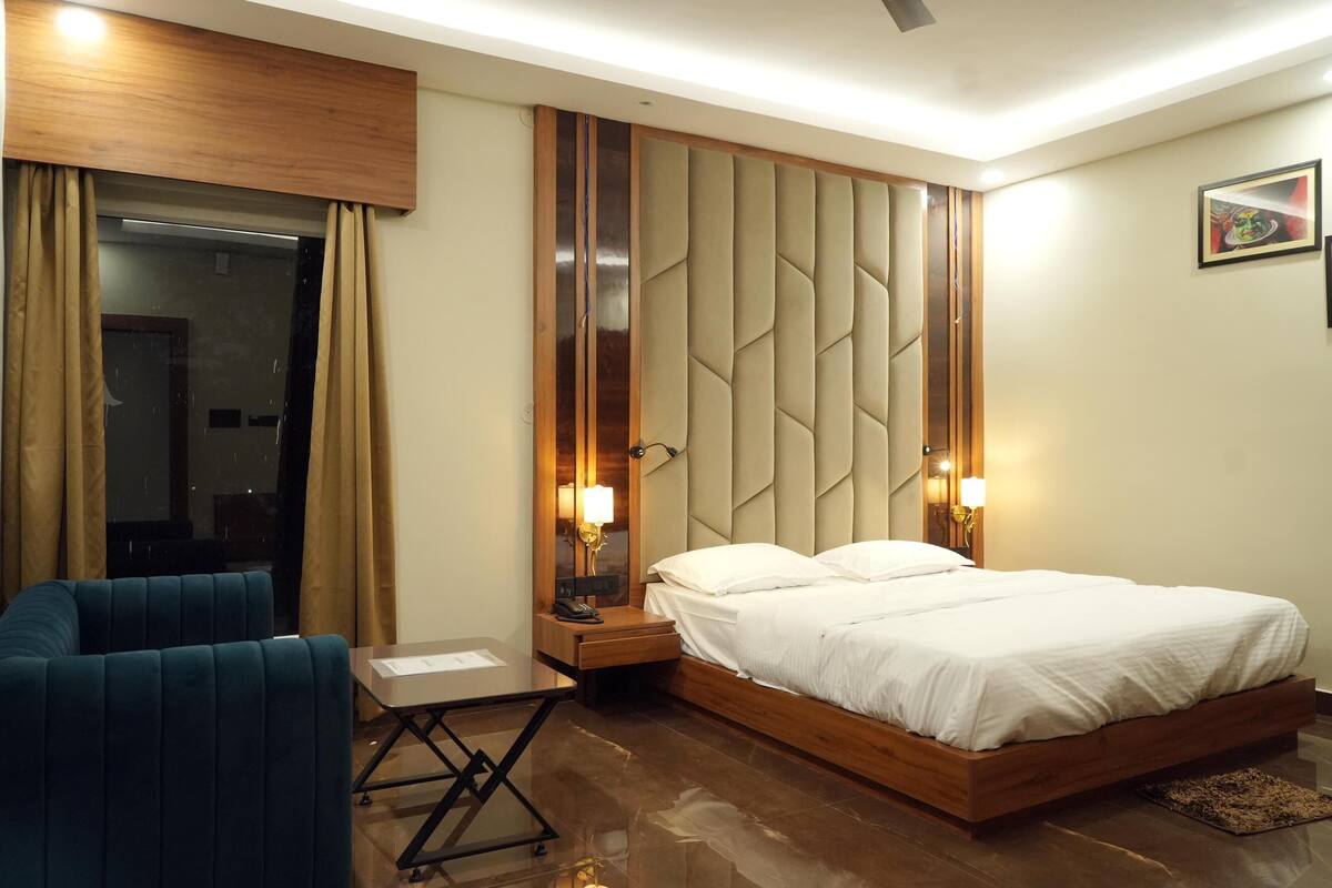 Una habitación de hotel moderna con cabecera de madera, cama con sábanas blancas, un sillón azul, una mesita pequeña y una mesita de noche con lámpara.