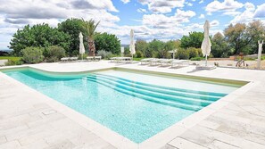 Ein rechteckiger Swimmingpool mit klarem, blauem Wasser, umgeben von weißen Liegestühlen und Sonnenschirmen, vor einer Kulisse aus Bäumen und wolkenlosem Himmel.