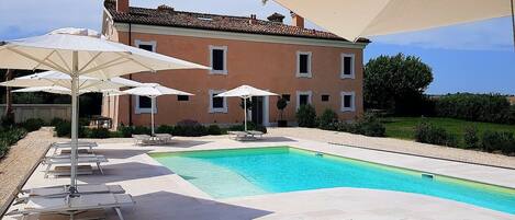 A swimming pool with lounge chairs and umbrellas, a large house in the background, and a well-maintained garden.